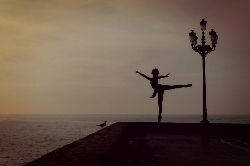 A women dancing at a pier by the water with a bird sitting next to her. The colours are neutral. The dance pose is strong and beautiful.
